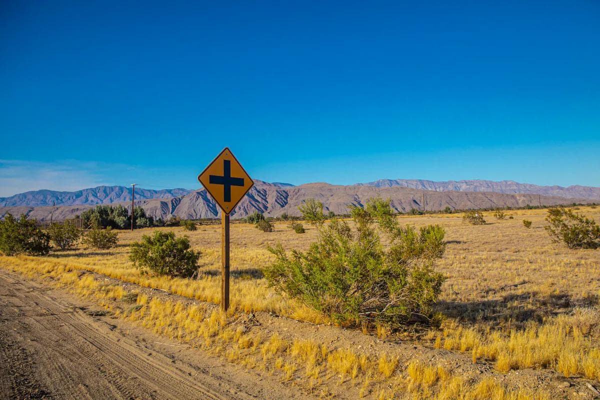 Yellow crossroad sign along a dirt road with mountains in distance.