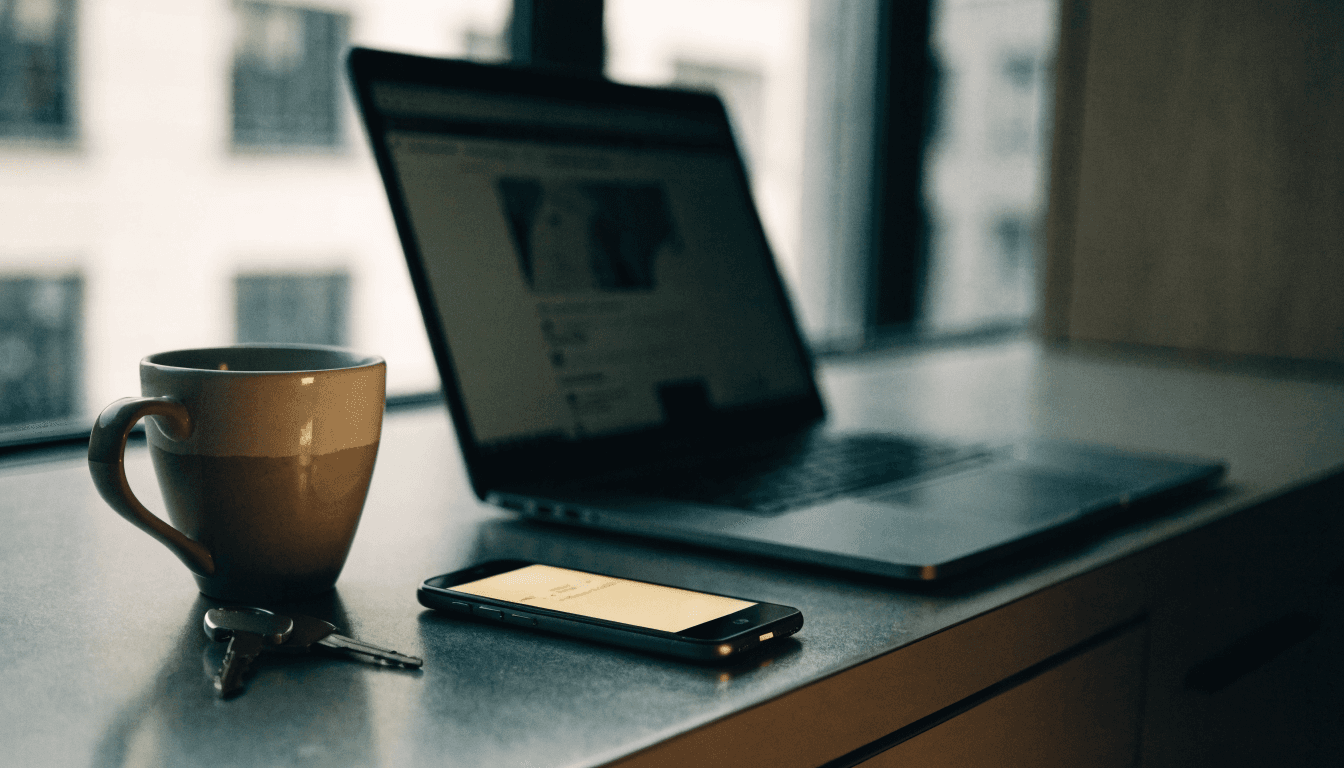 A laptop open on a kitchen worktop beside a mobile phone lit up mid-ring, with a mug and car keys nearby and nobody in frame